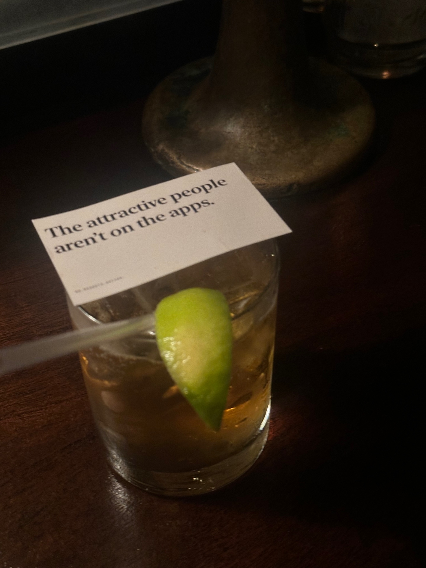 A card resting against a cocktail glass on a dark wooden bar. The card reads the attractive people aren't on the apps.